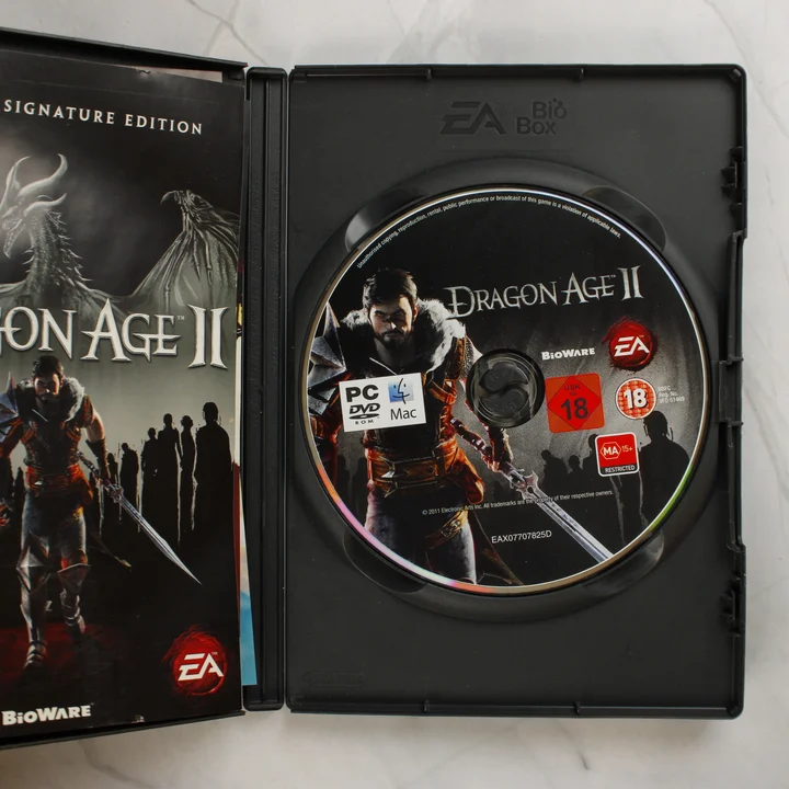Nyitott Dragon Age II videójáték tok a lemezzel és a füzettel