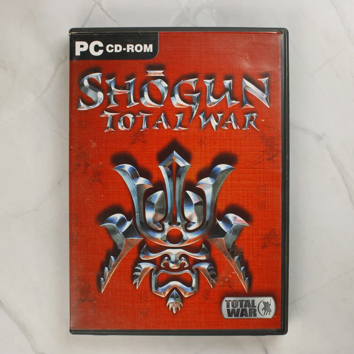 Shogun: Total War PC játék előlapja vörös háttérrel és szamuráj sisakkal.