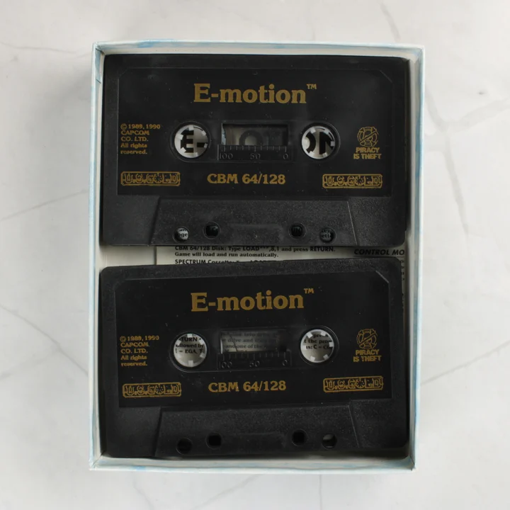 E-Motion kazetták Két fekete Commodore 64 kazetta az E-Motion játékhoz a dobozban
